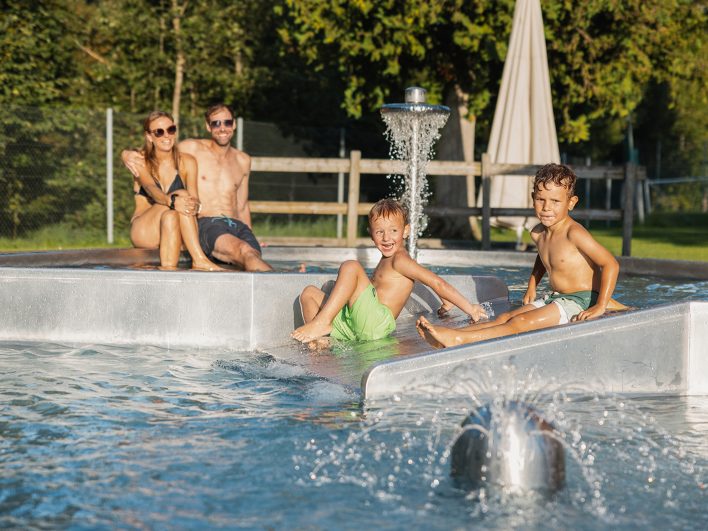 Zwei Jungen rutschen im Schwimmbad Hittisau ins Kinderbecken mit Wasserpilz, zwei Erwachsene sitzen lächelnd am Beckenrand in der Sonne.
