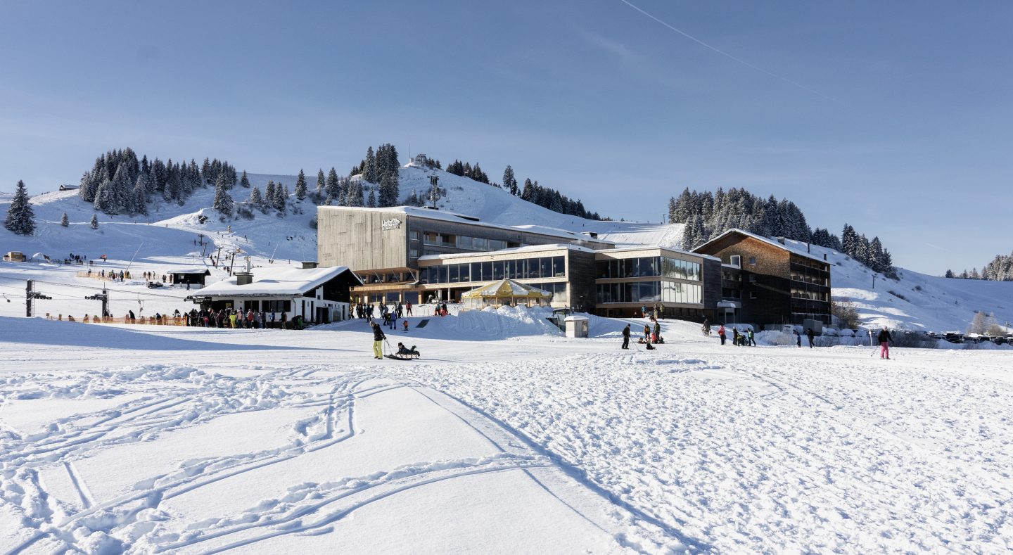 Almhotel Hochhäderich auf 1.300 Metern im Winter, direkt an den Pisten und Liften der Alpenarena Hochhäderich, davor zieht ein Vater einen Schlitten mit Kind über den Schnee.