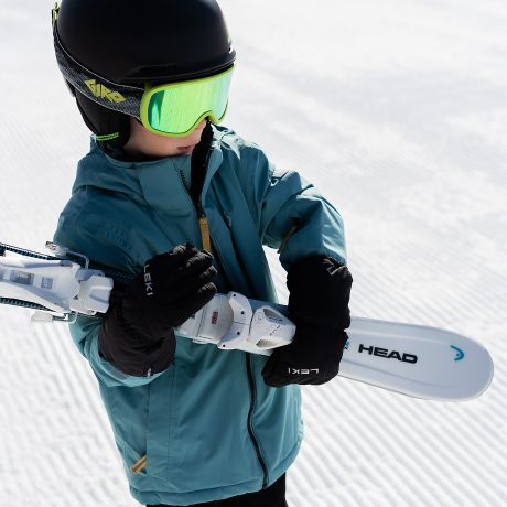 Kind mit Helm und neongelber Skibrille trägt stolz seine Ski über der Schulter, im Hintergrund die frisch präparierte Piste.