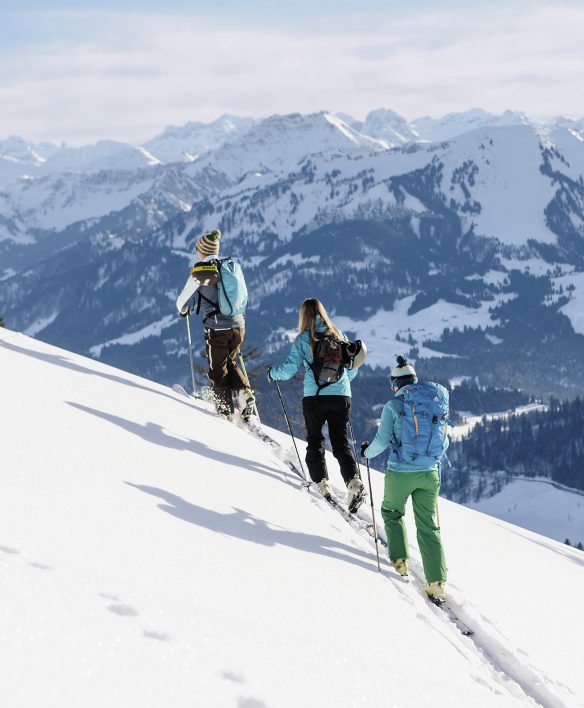 Drei Skitourengeherinnen spuren hintereinander einen steilen Schneehang hinauf, vor ihnen ein weites Panorama verschneiter Berge.