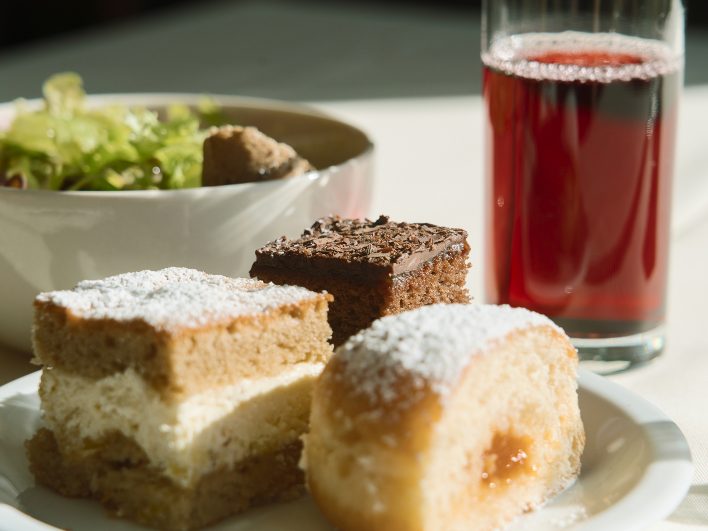 Süße Leckereien der Almjause im Almhotel Hochhäderich mit Schokoladenschnitte, Cremeschnitte und gefülltem Hefegebäck, dahinter eine Schale mit Salat und ein Glas roter Saft.
