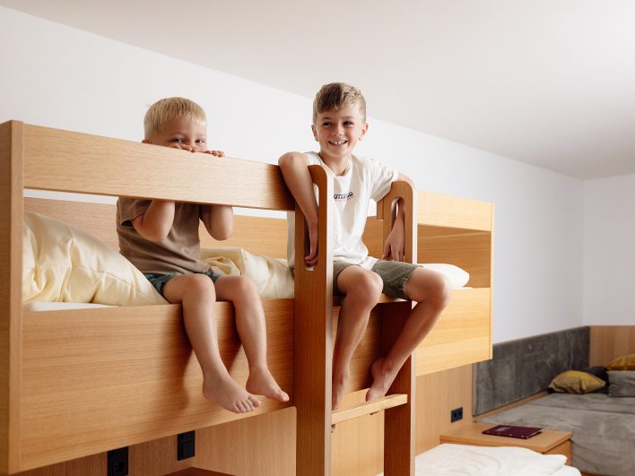 Zwei Jungen sitzen lachend auf dem oberen Stockbett aus hellem Holz im Familienzimmer, daneben eine Leiter zum Klettern.