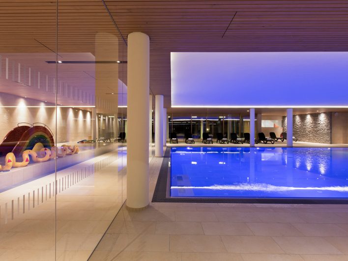 Panorama-Hallenbad im Almhotel Hochhäderich am Abend mit blau beleuchtetem Schwimmbecken und Liegen, links hinter einer Glaswand das Kinderbecken mit Regenbogenrutsche und Spritzschlange.