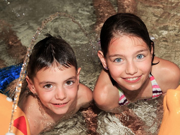 Ein Junge und ein Mädchen lächeln im Kinderbecken in die Kamera, während die Spritzschlange einen Wasserbogen über sie sprüht.