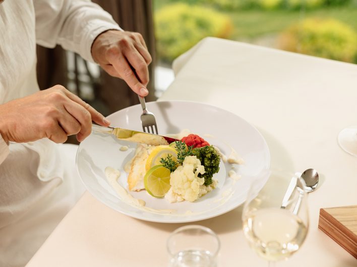 Gast schneidet beim Abendessen ein Fischfilet mit Blumenkohl, Brokkoli und Zitrone an, daneben ein Glas Weißwein.