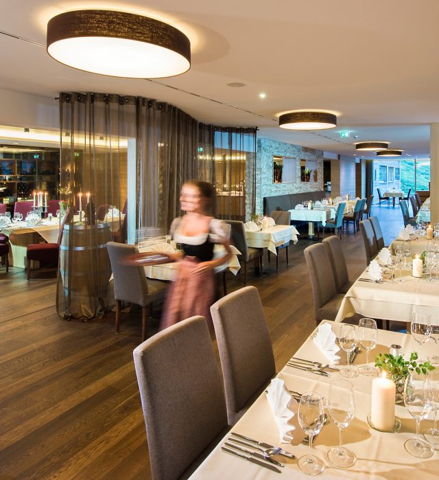 Hotelrestaurant im Almhotel Hochhäderich mit festlich eingedeckten Tischen, Kerzenlicht und großen Fenstern, eine Mitarbeiterin im Dirndl bringt lächelnd ein Tablett an den Tisch.