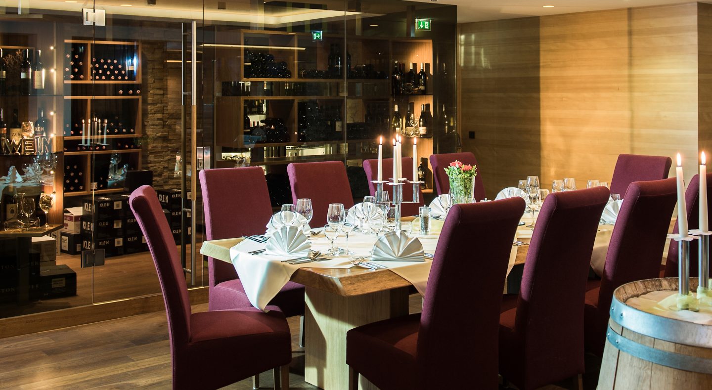 Gedeckte Tafel mit Kerzenleuchtern und weinroten Stühlen an einem massiven Holztisch vor der gläsernen Vinothek des Almhotels Hochhäderich, in deren Regalen zahlreiche Weinflaschen lagern.