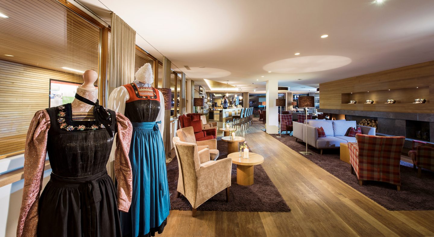 Lounge der Wälderbar im Almhotel Hochhäderich mit Sesseln, Sofas und Kaminfeuer, vorne zwei traditionelle Bregenzerwälder Trachten auf Schneiderpuppen.