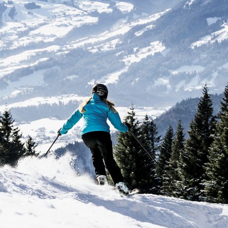Skifahrerin in türkiser Jacke schwingt eine Piste hinunter, unter ihr öffnet sich der weite Blick auf verschneite Täler und Dörfer im Bregenzerwald.