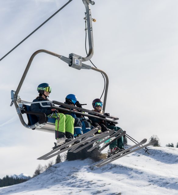 Drei lachende Skifahrer mit Helm und bunter Skibrille fahren im kindergesicherten Vierersessellift der Alpenarena Hochhäderich bergauf.