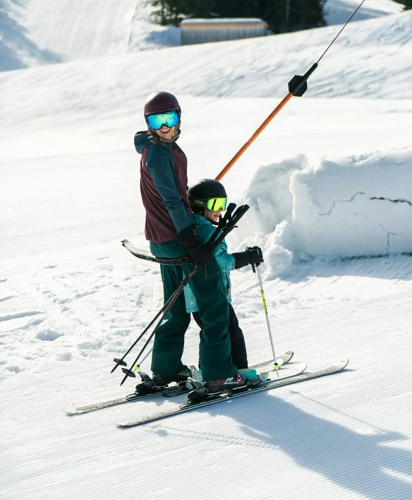 Erwachsene und Kind fahren gemeinsam mit dem Schlepplift den Übungshang hinauf, die Frau lacht dabei in die Kamera.