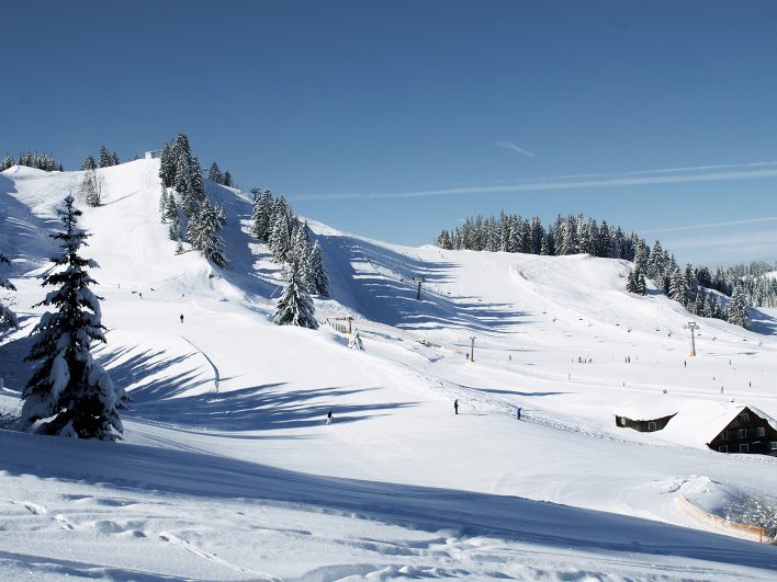 Breite, sonnige Skipisten der Alpenarena Hochhäderich mit Sessellift, verschneiten Fichten und einer alten Holzhütte am Pistenrand.