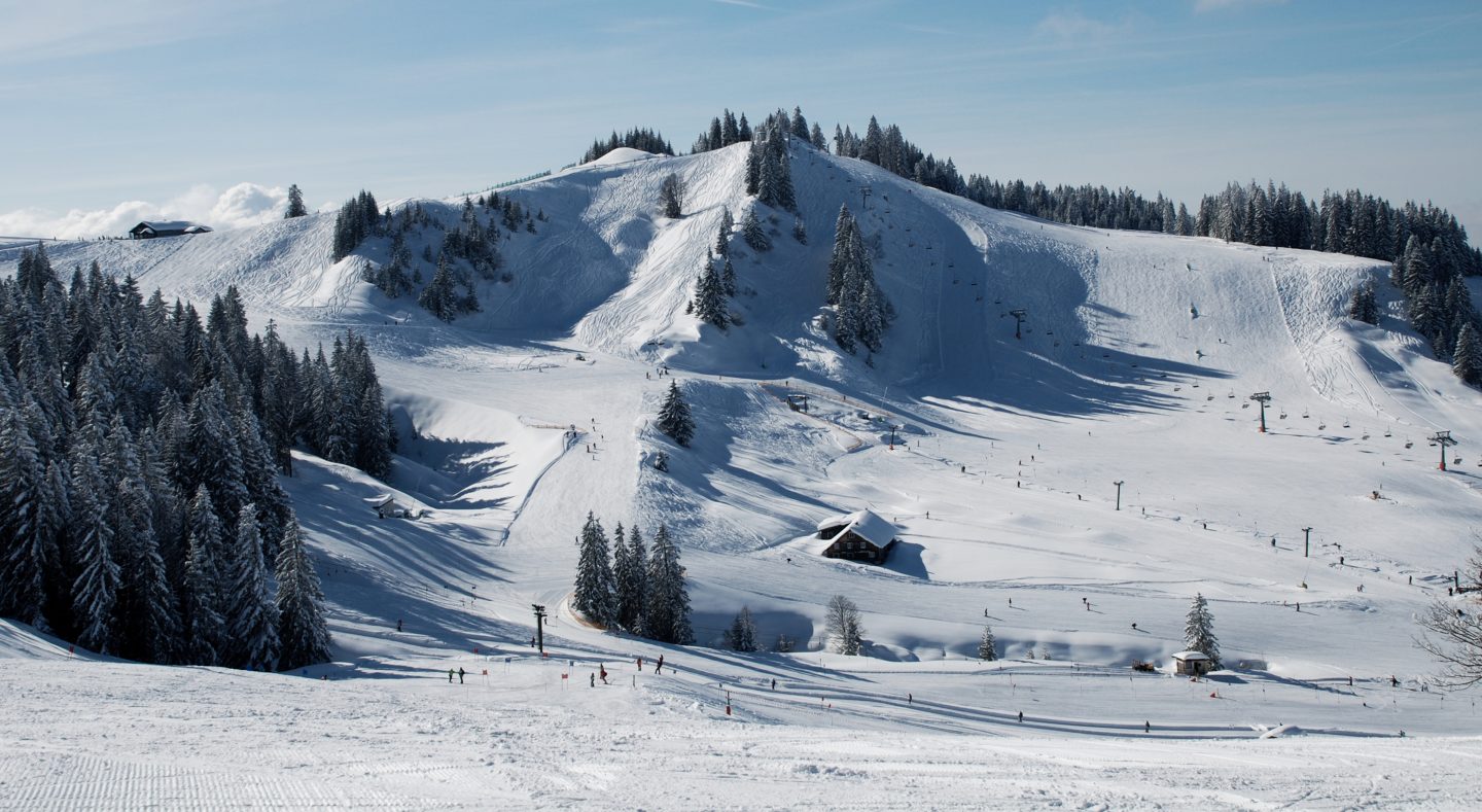 Übersicht über das Skigebiet Alpenarena Hochhäderich zwischen 1.300 und 1.600 Metern mit Sessellift, breiten Pisten, verschneiten Hütten und Fichtenwald unter blauem Himmel.