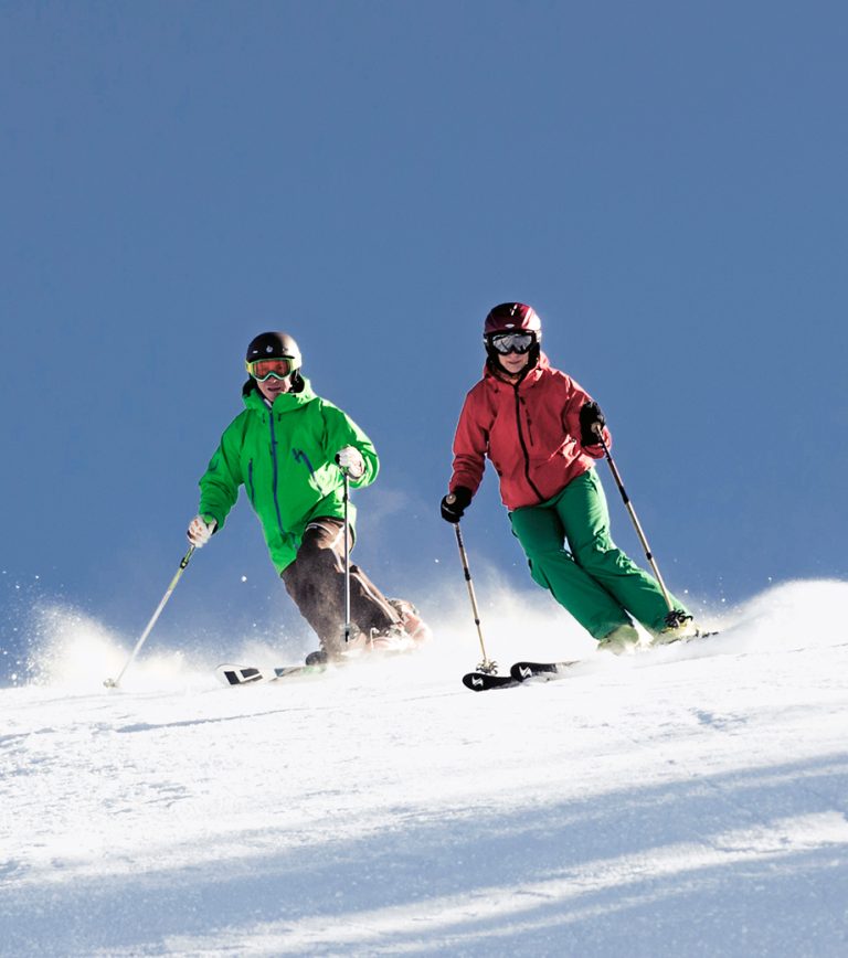Zwei Skifahrerinnen in grüner und roter Jacke ziehen bei blauem Himmel ihre Schwünge über die Piste.