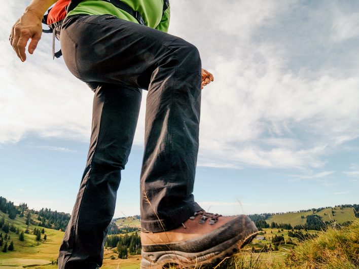 Wanderer mit festen Bergschuhen setzt zum nächsten Schritt an, darunter weite Almwiesen und Fichtenwälder im Bregenzerwald unter blauem Himmel.