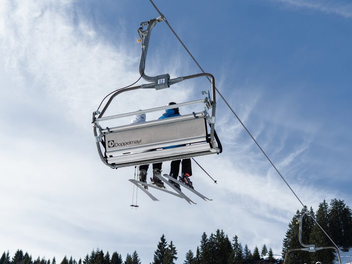 Zwei Skifahrer schweben im Sessellift der Alpenarena Hochhäderich über verschneiten Fichten nach oben, dahinter blauer Himmel mit zarten Schleierwolken.