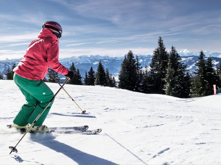 Skifahrerin in roter Jacke fährt über eine sonnige Piste, dahinter reicht der Blick über Fichten bis zu den verschneiten Bergketten der Alpen.