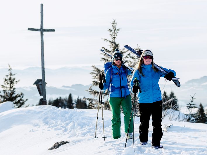 Zwei gut gelaunte Skitourengeherinnen tragen ihre Ski geschultert über einen verschneiten Gipfel, hinter ihnen ein hölzernes Gipfelkreuz.