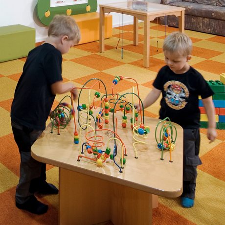 Zwei kleine Jungen schieben bunte Holzperlen über die Drahtschleifen eines Motorikspieltischs, im Hintergrund große Bausteine und Sitzwürfel im Kleinkindbereich.