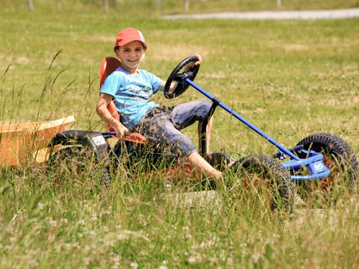 …_Spielealm_Gokart.jpg Fröhlicher Junge mit Schirmmütze fährt im Tretauto mit Anhänger durch die sommerliche Almwiese auf der Spielealm.