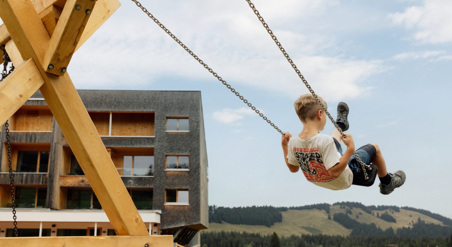 Junge schaukelt hoch hinaus auf der Spielealm, im Hintergrund die Holzfassade des Almhotels und bewaldete Almhänge.