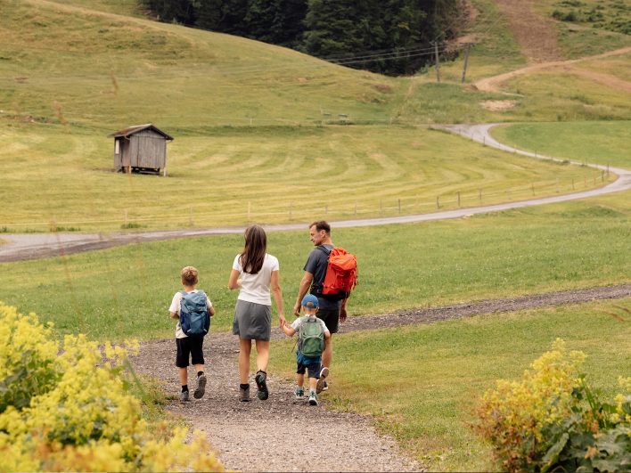 Familie mit Kinderrucksäcken wandert von hinten gesehen auf einem Kiesweg über Almwiesen, vorbei an einer alten Holzhütte.