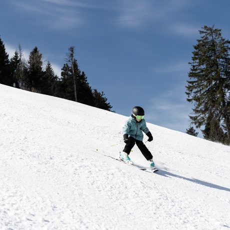 Kind mit Helm und Skibrille fährt eine sonnige Piste der Alpenarena Hochhäderich hinunter, gesäumt von verschneiten Fichten unter blauem Himmel.