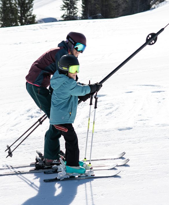 Kind und erwachsene Begleitung lassen sich gemeinsam am Schlepplift den flachen Übungshang hinaufziehen, beide mit Helm und bunter Skibrille.