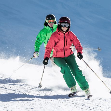 Paar in bunten Skijacken fährt hintereinander eine sonnige Skipiste im Bregenzerwald hinunter, der Schnee stiebt bei jedem Schwung.