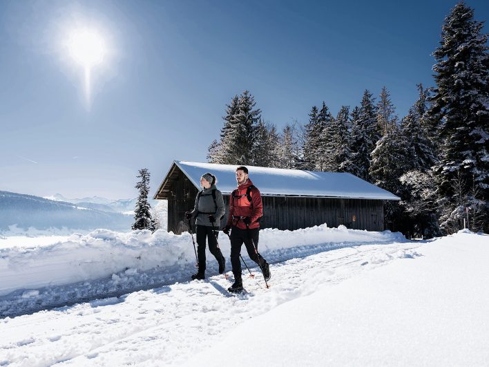 Paar bei einer Winterwanderung auf einem verschneiten Weg im Bregenzerwald, vorbei an einem alten Holzstadel und tief verschneiten Fichten im strahlenden Sonnenschein.
