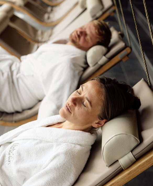 Paar in weißen Bademänteln mit Hotellogo ruht mit geschlossenen Augen nebeneinander auf Schwebeliegen im Wellnessbereich.