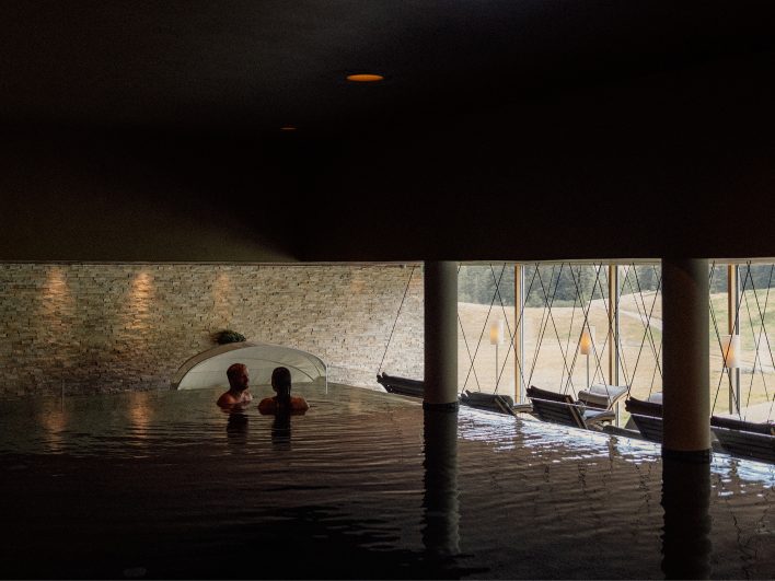 Paar im abgedunkelten Schwimmbecken des AlmSpa, dahinter eine beleuchtete Natursteinwand und die Fensterfront.