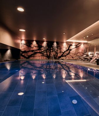 Nacktschwimmbecken im Adults-only AlmSpa des Almhotels Hochhäderich mit einer Frau auf einer Liege am blau beleuchteten Pool, die Zeitung ließt.