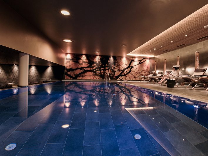 Nacktschwimmbecken im Adults-only AlmSpa des Almhotels Hochhäderich mit einer Frau auf einer Liege am blau beleuchteten Pool, die Zeitung ließt.