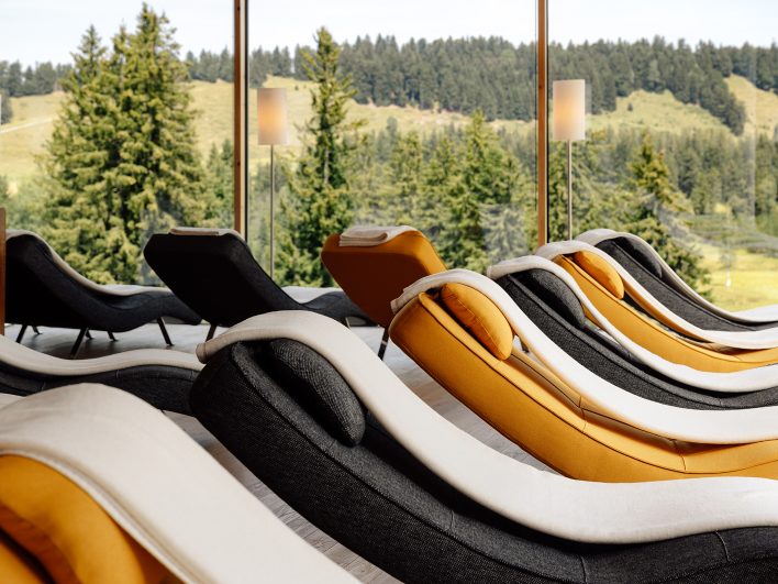 Reihe geschwungener Relaxliegen in Anthrazit, Creme und Orange vor der Fensterfront des Ruheraums mit Blick auf Fichten.