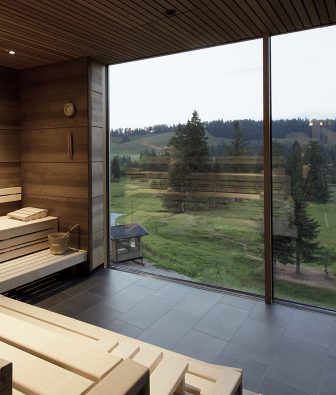 Finnische Sauna im AlmSpa mit hellen Holzbänken und raumhoher Glasfront, draußen Almwiesen, Fichten und eine kleine Holzhütte im Abendlicht.