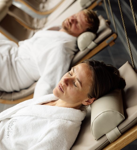 Frau und Mann in weißen Bademänteln mit Hotellogo ruhen mit geschlossenen Augen auf Schwebeliegen im Day Spa.