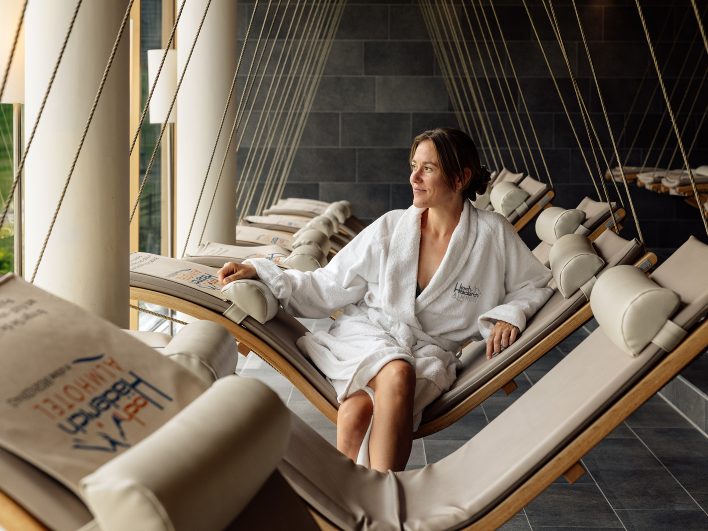 Frau im Bademantel sitzt auf einer Schwebeliege im Alm Spa und schaut glücklich nach draußen.