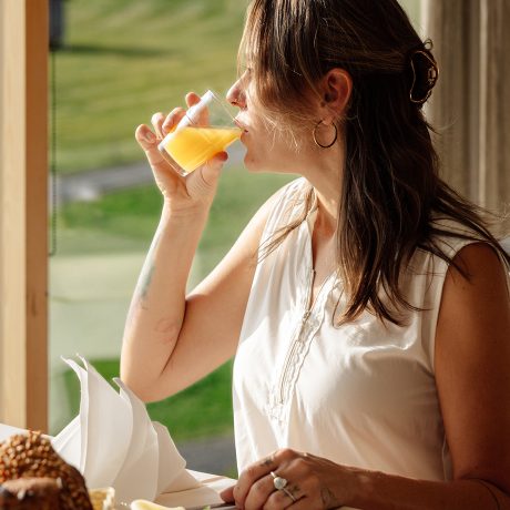 Frau trinkt beim Frühstück im Almhotel Hochhäderich ein Glas Orangensaft, durch das Fenster leuchtet eine grüne Almwiese.