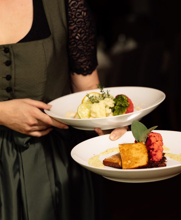 Servicemitarbeiterin im grünen Dirndl trägt zwei Teller mit bunt angerichtetem Blumenkohl, Kräutern und Sauce an den Tisch.