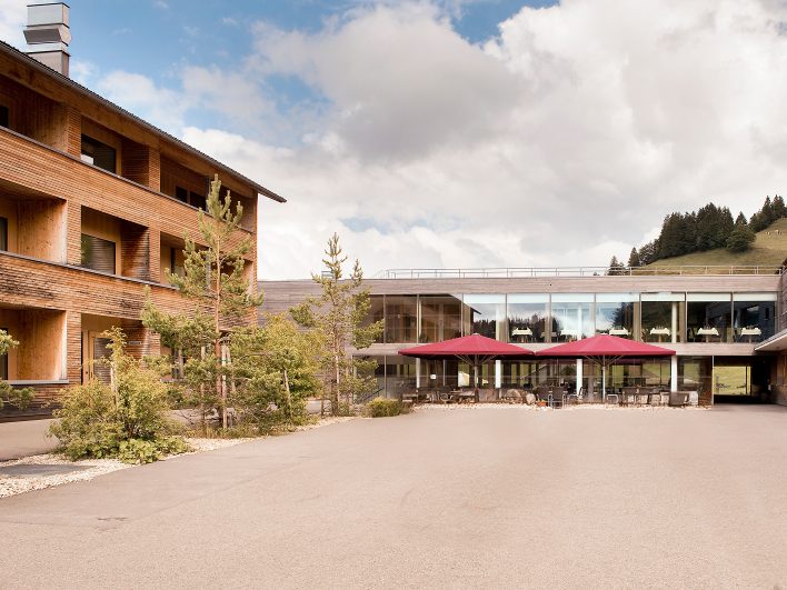 Innenhof des Almhotel Hochhäderich mit Holzfassade und verglastem Verbindungsbau, davor eine Terrasse mit roten Sonnenschirmen am bewaldeten Hang.