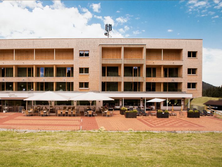 Holzschindelfassade des Almhotel Hochhäderich mit dem Tagesrestaurant Johann & Frieda im Erdgeschoss und großer Sonnenterrasse mit Sonnenschirmen und Liegen.