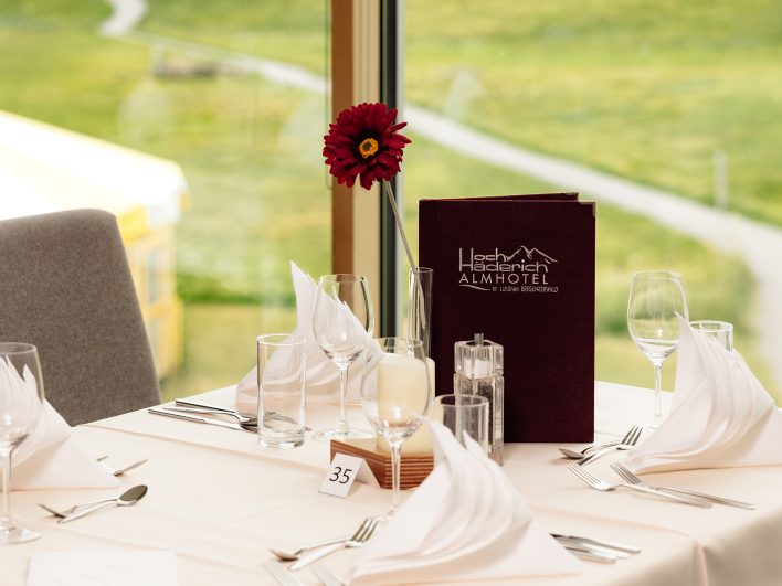 Festlich gedeckter Tisch mit Weingläsern, weißen Servietten, roter Gerbera und Getränkekarte des Almhotel Hochhäderich im Bregenzerwald vor einem großen Fenster.