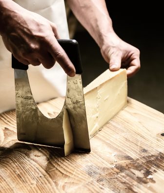 Hände schneiden mit einem großen Käsemesser ein Stück gereiften Hartkäse auf einem Holzbrett