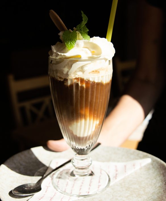 Eiskaffee im hohen Glas mit Vanilleeis, Schlagsahne, Minzblatt und Strohhalm, serviert auf einem Teller mit Löffel.