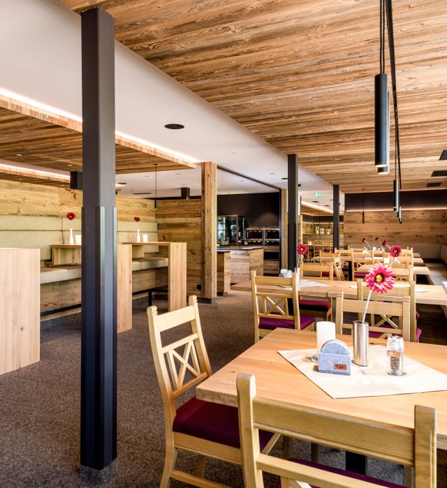 Gastraum des Johann & Frieda am Almhotel Hochhäderich mit Altholzdecke, gemütlichen Sitznischen und Holzstühlen mit roten Sitzpolstern, auf jedem Tisch eine rote Blume.