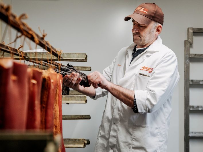 Metzger Anton Fetz prüft in der Reifekammer ein Stück geräuchertes Fleisch, davor hängen Speckseiten zum Trocknen.