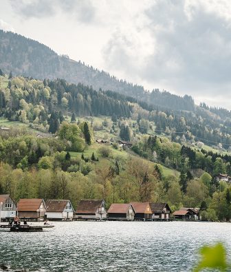 Bootshäuser am Ufer des Alpsees bei Immenstadt im Allgäu, dahinter bewaldete Hänge mit Wiesen und Höfen