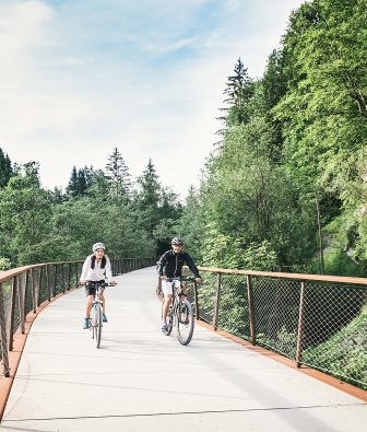 Zwei Radfahrer mit Helm auf einer Brücke des Radwegs im Bregenzerwald, umgeben von sommergrünem Wald