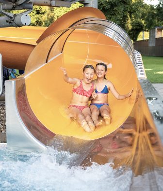Zwei lachende Mädchen rutschen gemeinsam aus einer orangen Wasserrutsche ins Becken des Schwimmbads Hittisau.
